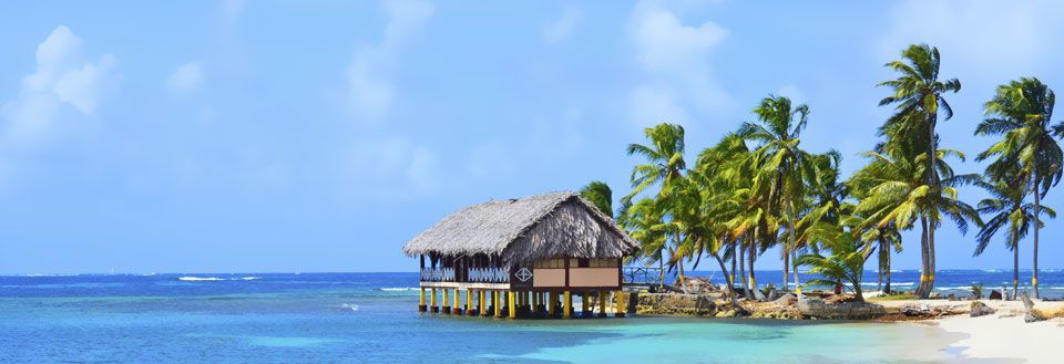 En stråtekket hytte over vannet omringet av palmer på en tropisk strand.