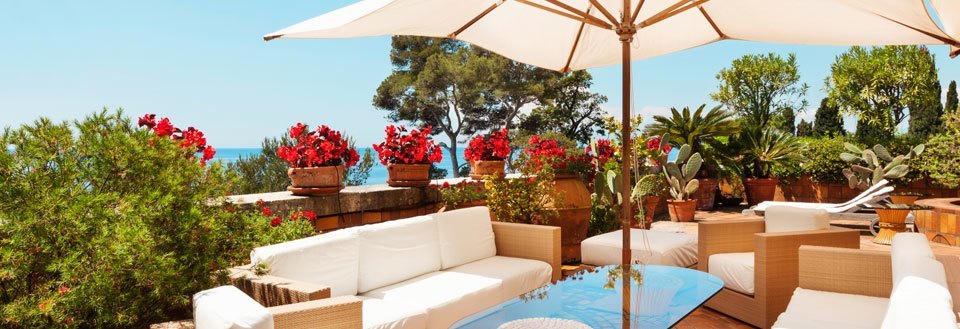 En koselig terrasse med hvite sofaer, en parasoll, frodige planter og potteblomster under den blå himmelen.
