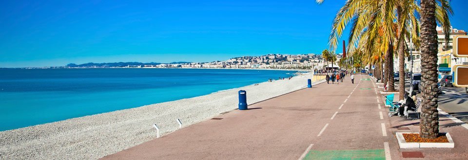 En solfylt promenade i Nice med palmetrær langs en klar blå kystlinje med en by i det fjerne.