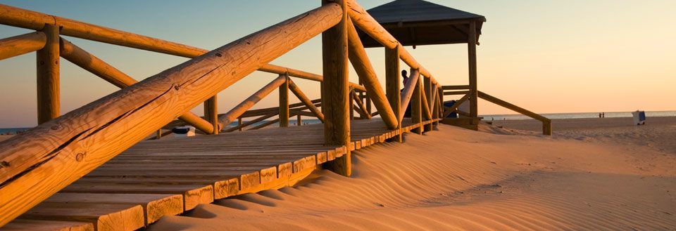 En trebro strekker seg over sanddynene mot en strand under solnedgangen under en klar himmel.