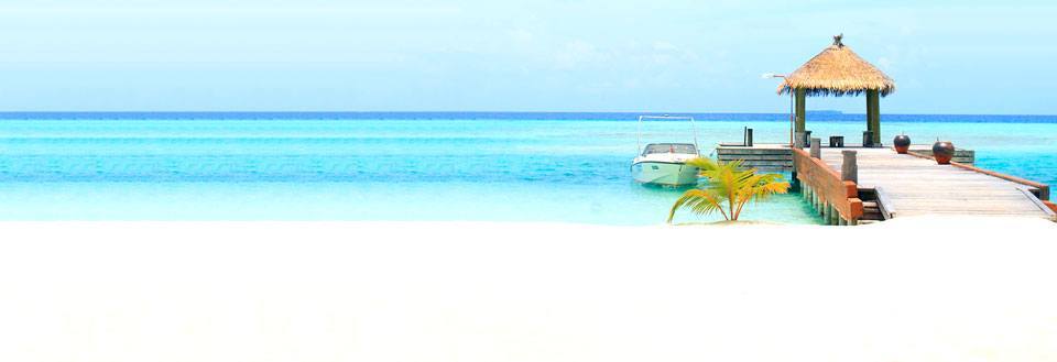 Et fredfullt strandbilde med hvit sand, en trebrygge som strekker seg mot en hytte med stråtak og en båt.