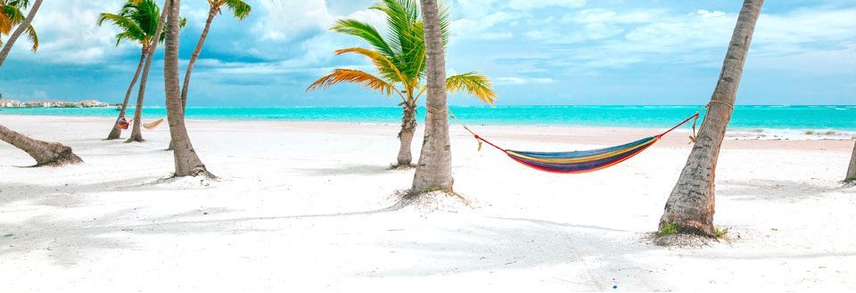 En hengekøye mellom palmetrær på en hvit sandstrand med klar blå himmel og hav i bakgrunnen.