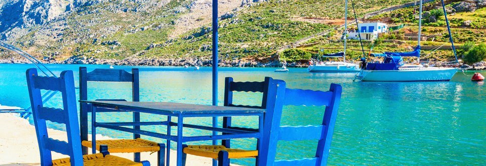 Et fredelig strandbilde med blå stoler og bord foran en krystallklar blå bukt med båter og fjell i bakgrunnen.