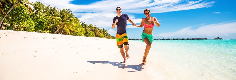En mann og en kvinne smiler og løper langs en solfylt strand med palmer i bakgrunnen.