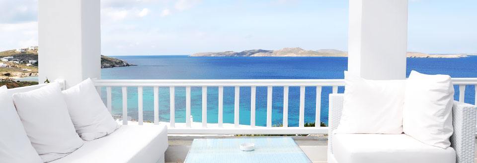 Bildet viser en terrasse med hvit sofa og puter, med utsikt over et klart blått hav og himmel.