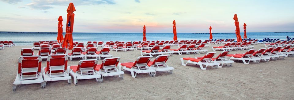 Tomme røde solsenger på en sandstrand med lukkede parasoller, havet i bakgrunnen under skumring.