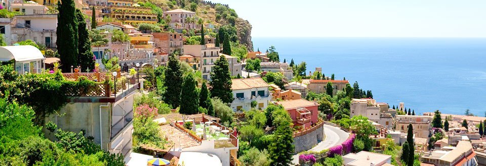 Bilde av en idyllisk kystby med fargerike hus og frodig vegetasjon mot en klar blå himmel.