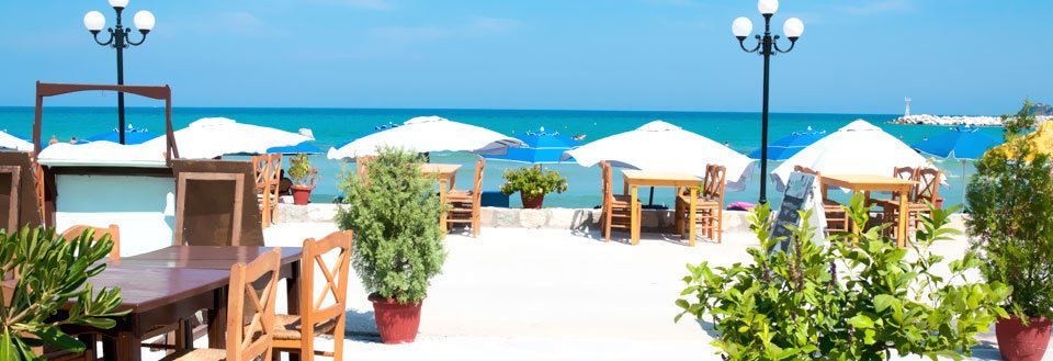 En sjarmerende strandrestaurant med trebord og stoler under hvite parasoller, med havet i bakgrunnen.