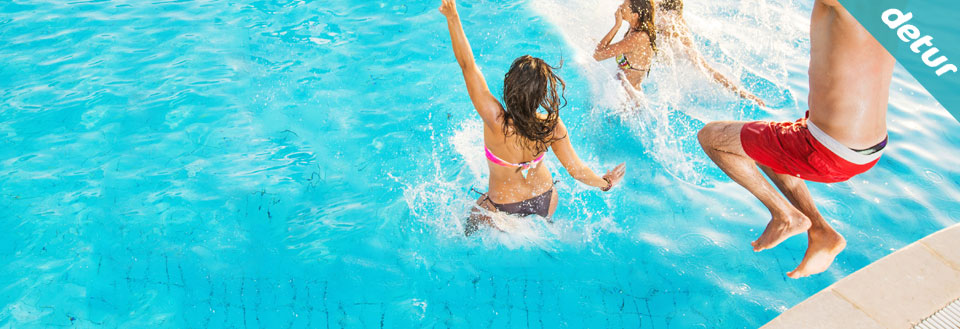 To mennesker har det gøy med å hoppe inn i en klar, blå svømmebasseng i solskinn.