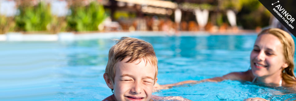 En ung gutt smiler bredt mens han svømmer ved siden av en smilende kvinne i et utendørs svømmebasseng.