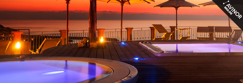 Utsikt over et opplyst svømmebasseng og terrasse ved solnedgang med havutsikt.