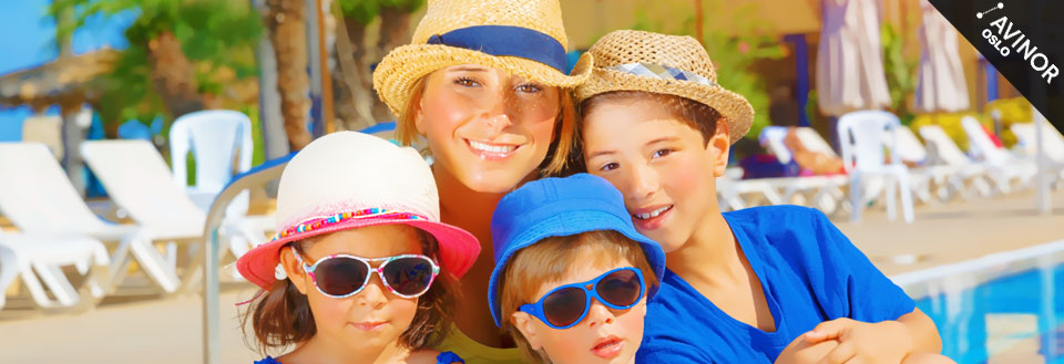 En kvinne og tre barn smiler mot kameraet med sommerhatter og solbriller ved en svømmebasseng.