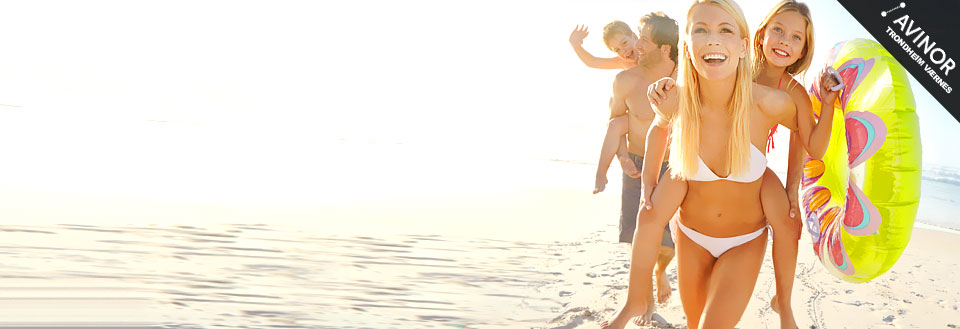 En familie koser seg i solen på stranda; barna leker, og en kvinne smiler med badetøy og badebøye.