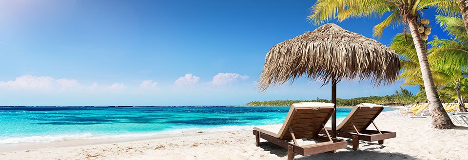 To solsenger under en stråparasoll på en hvit sandstrand med krystallklart blått hav og palmetrær.