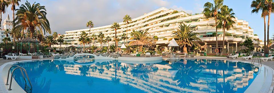 Bildet fremviser et fleretasjes luksushotell omgitt av palmer og et stort svømmebasseng, badet i morgenlyset.