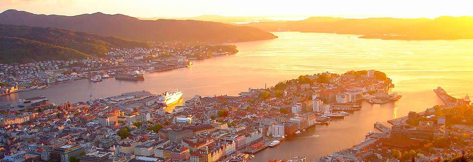Bilde av en by ved solnedgang med fjell i bakgrunnen og en glitrende elv eller fjord i forgrunnen.