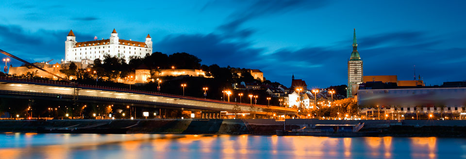 Et kveldsbilde av Donau i Bratislava med en bro i forgrunnen og en belyst borg på en høyde i bakgrunnen.