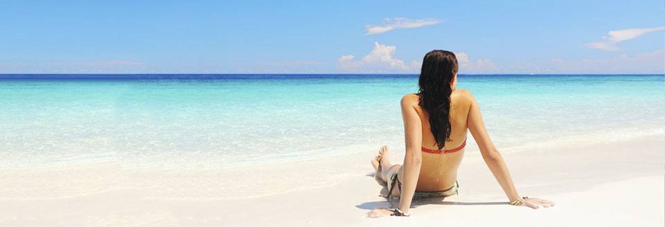 En person sitter på en hvit sandstrand og ser utover det asurblå havet.