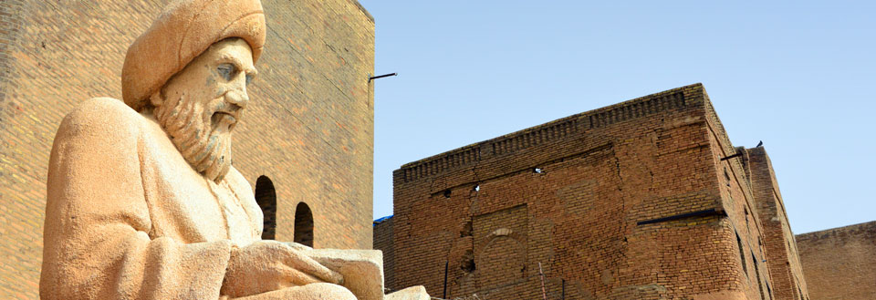 Bildet fremviser en sandsteinstatue av en skjeggete mann med turban foran en mursteinsbygning under en klar blå himmel.