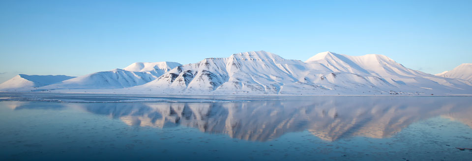 Speilblankt vann med tydelige refleksjoner av snødekte fjell og en klar blå himmel.