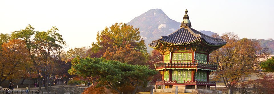 Et tradisjonelt asiatisk palass i Seoul omringet av gyldne høsttrær med fjell i bakgrunnen.