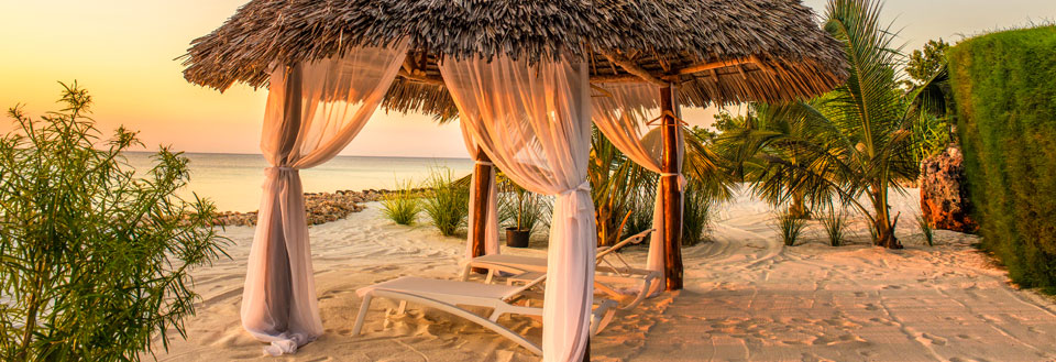 Romantisk strandhytte med hvite gardiner i solnedgangen, klar for avslapning.