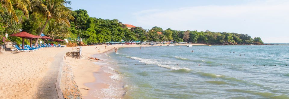 Strandlandskapet fremviser en solfylt dag med mennesker som nyter sjøen, sandet og strandparasollene under tropiske trær.