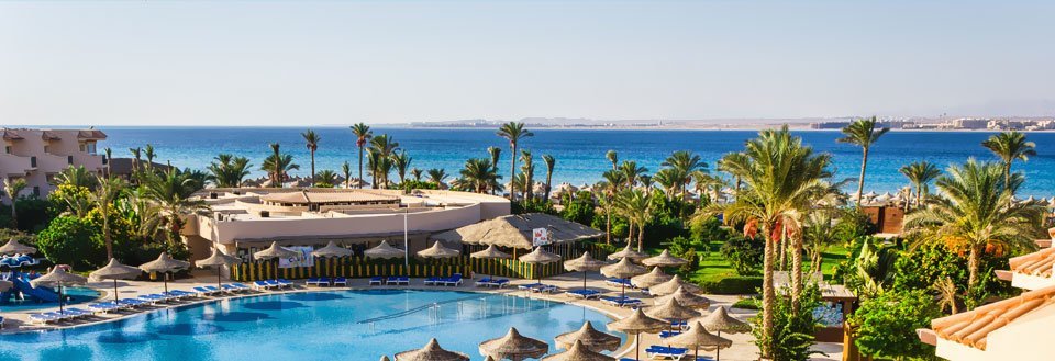 Et feriested med svømmebasseng, palmer og utsikt mot stranden under en klar himmel.