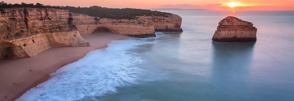 Solnedgangen kaster myke farger over en strand med steinete formasjoner i vannet.