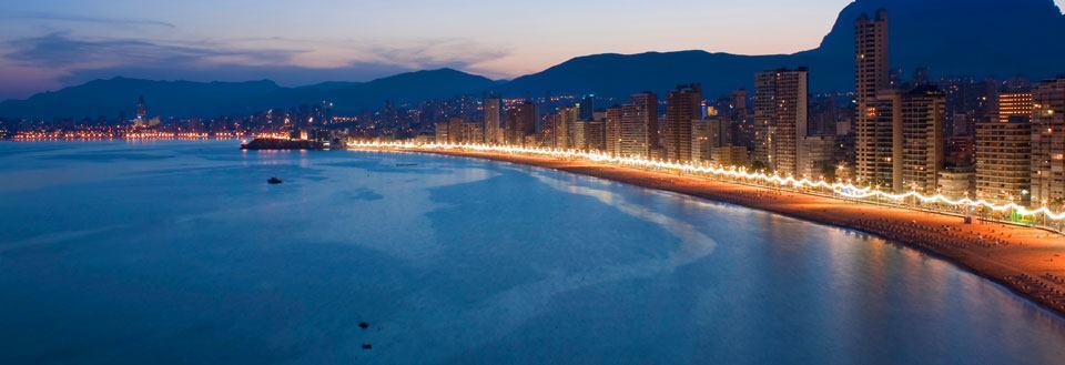 En by ved kysten lyser opp om kvelden, med en lang strand og skyskrapere.
