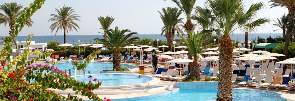 Luksuriøs ferieresort med svømmebasseng, palmer og parasoller langs en solrik kyst.