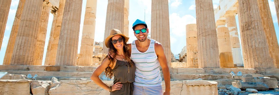 To glade mennesker poserer foran antikke søyler under en klar himmel i Athen.