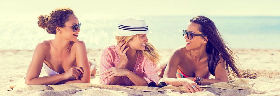 Tre venninner ler og prater på en solfylt strand med blå himmel og havet i bakgrunnen.