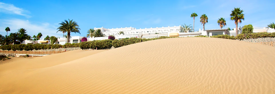Solfylt ørken med sanddyne, hvite bygninger og palmer i bakgrunnen.