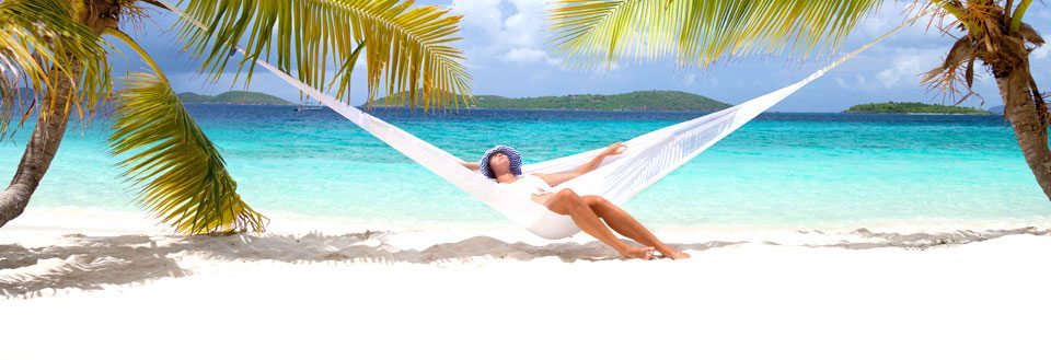 En person hviler i en hengekøye mellom to palmer ved en tropisk strand med klart blått vann.