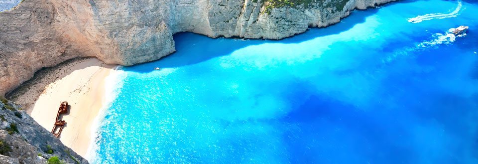 En idyllisk strand omgitt av klipper med krystallklart blått vann. Et skip seiler i nærheten.