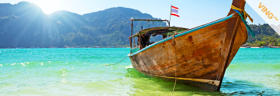 En tradisjonell båt flyter på krystallklart vann med fjell i bakgrunnen og solskinn.