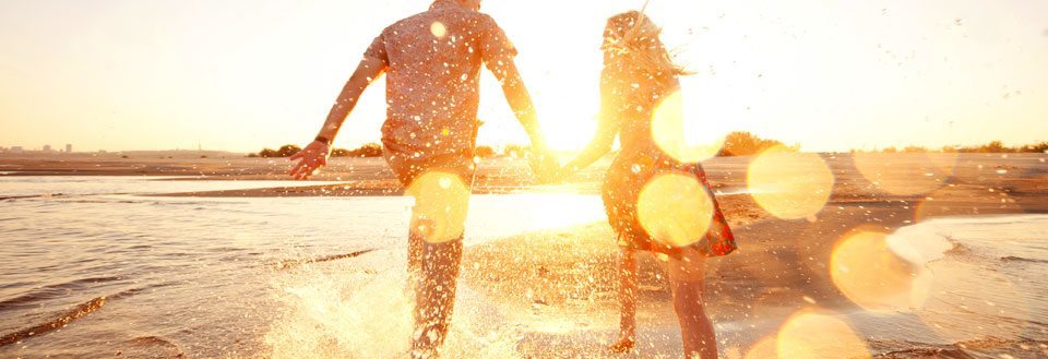 To personer hånd i hånd løper langs vannkanten på en strand i solnedgangens skjær.