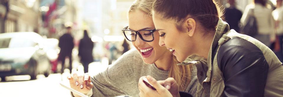 To smilende kvinner ser på en smarttelefon sammen på en travel bygate.