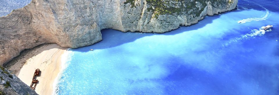 Storartet strand med høye klipper og krystallklart blått vann. En båd seiler i nærheten.
