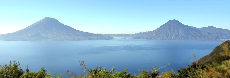 Panoramautsikt over en rolig innsjø flankert av fjell under en klar blå himmel.
