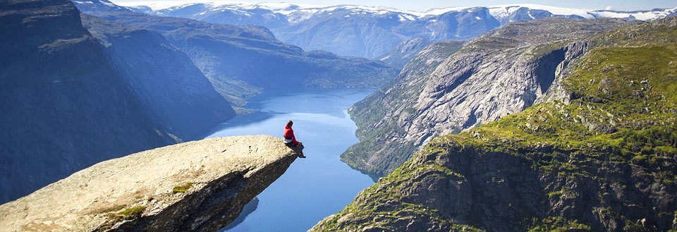 En person sitter på kanten av et fjellutspring med utsikt over en fjord og fjell.