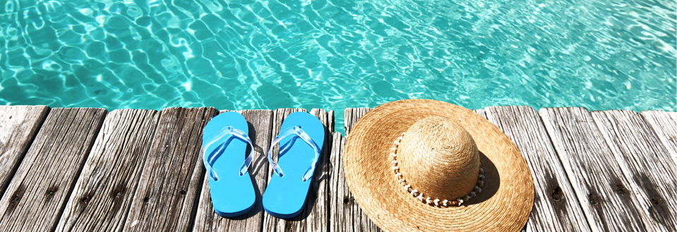 Blå flip-flops og en stråhatt hviler på en trebrygge ved siden av et svømmebasseng.