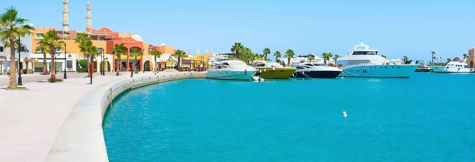 Bilde av en solrik havnepromenade  i Hurghada med luksusyachter fortøyd langs kaia foran fargerike bygninger.