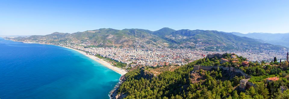 Panoramautsikt over Antalya med blått vann, strender og grønnkledde fjell i bakgrunnen.