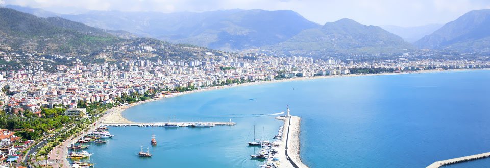 Panoramautsikt over Alanya med klart blått hav, en molo, båter og omringet av fjell.