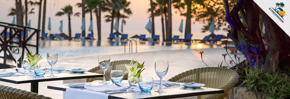 Et utendørs spiseområde ved svømmebassenget med solnedgang, palmer og solsenger i bakgrunnen.