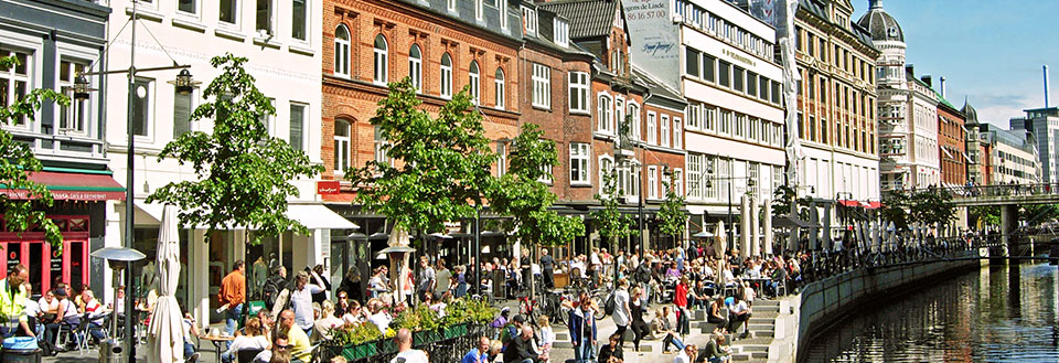 Glade mennesker nyder livet ved åen i Aarhus, en bro og grønne trær under en solfylt himmel.