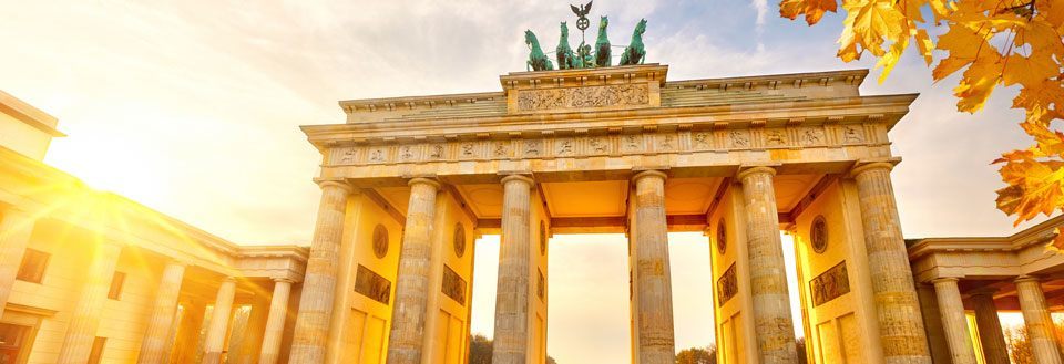 Bildet fremviser Brandenburger Tor i Berlin under et gyldent solnedgangslys, med høstløv foran.