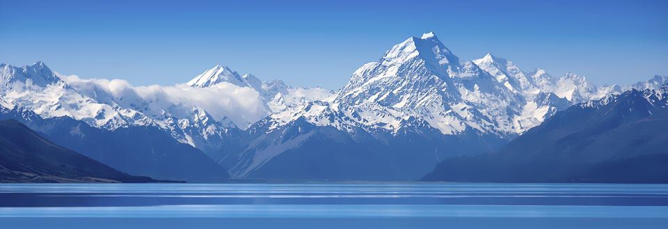 Panorama av en glitrende innsjø i Mount Cook nasjonalpark  foran majestetiske, snødekte fjell under en klar blå himmel.